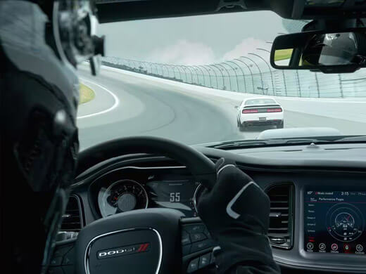 The interior of a 2023 Dodge Challenger as the driver takes a curve on a track, with the Digital Cluster displaying 55 mph and the Uconnect touchscreen displaying a Dodge Performance Page.