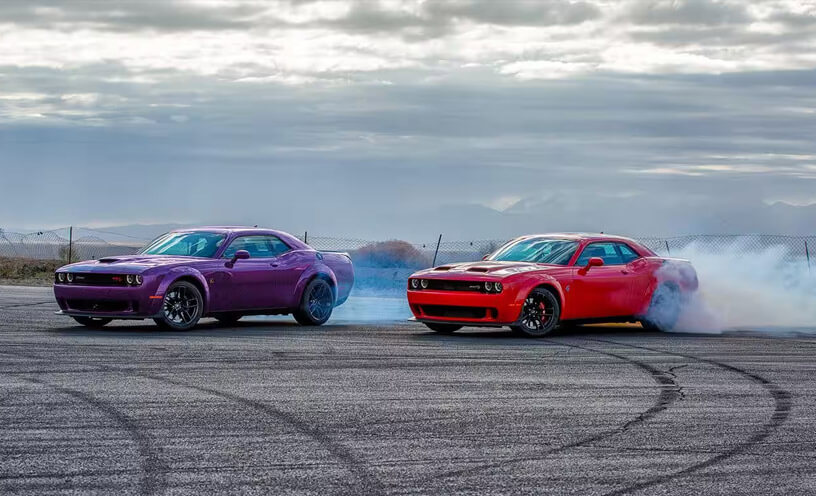 Two Dodge Challenger models making donuts on blacktop, a purple 2023 Challenger R/T and a red 2023 Challenger SRT Hellcat.