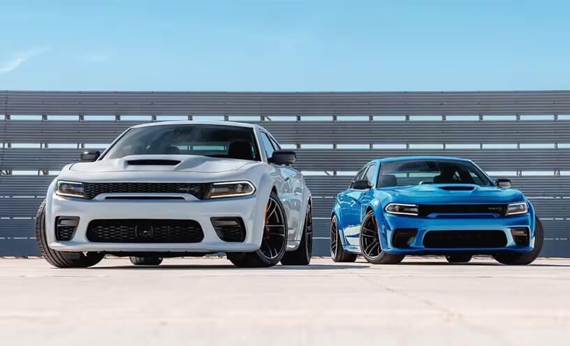 Two Dodge Charger models parked at a track. On the left: a white 2023 Dodge Charger SRT Hellcat. On the right: a blue 2023 Dodge Charger SRT Hellcat.